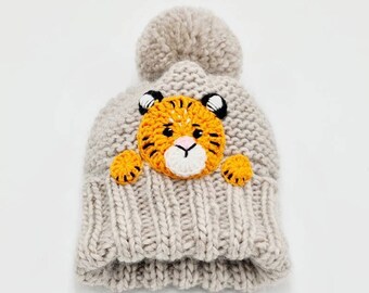May include: A light gray knit beanie with a large pom-pom on top. The front of the hat features a crocheted tiger face with orange and black stripes, black ears, and a white muzzle. The hat is made of a chunky knit material.