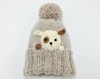 May include: A light beige knitted beanie with a matching pom-pom on top. The hat features a cream-colored dog face with brown accents, including a spot, ear, and nose. The brim is ribbed.