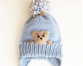 May include: A light blue knitted baby hat with a brown bear peeking out from the front. The hat has a white and brown pom pom on top and long ties with white and brown stripes.