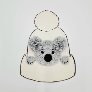 Könnte beinhalten: Eine weiße Strickmütze mit einem grauen gehäkelten Koalabild auf der Vorderseite. Die Mütze hat einen weißen Bommel oben.