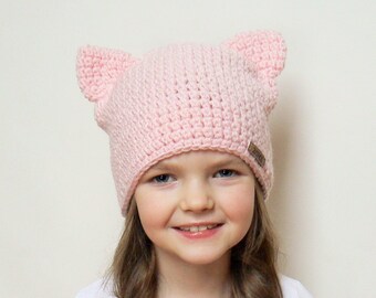 May include: A young girl wearing a pink crocheted hat with cat ears. The hat has a brown label with the word "Little" on it.
