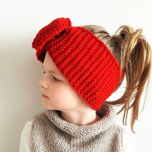 May include: A red crocheted headband with a large bow on top. The headband is worn by a young person with their hair pulled back in a ponytail.