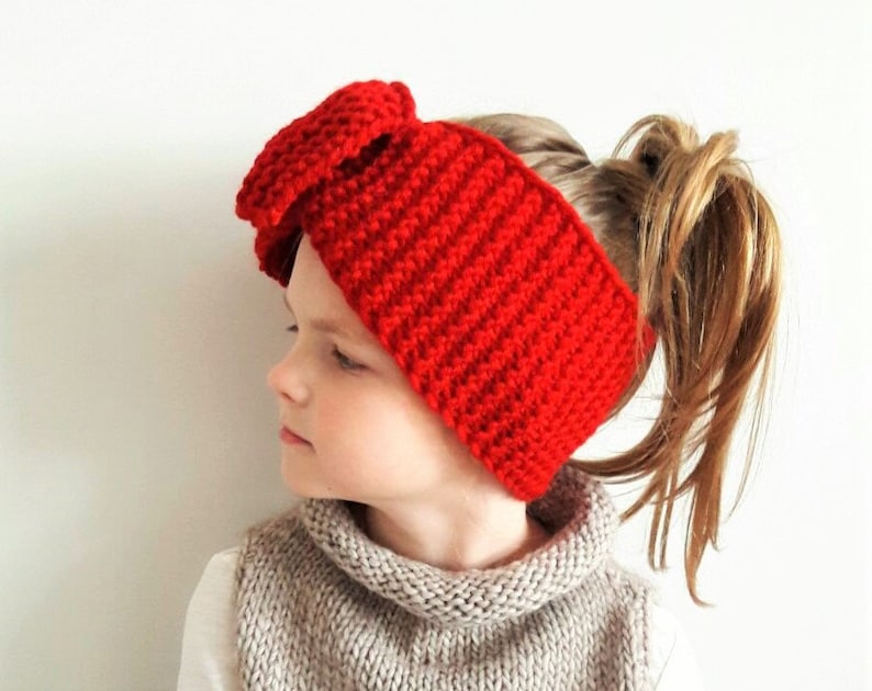 May include: A red crocheted headband with a large bow on top. The headband is worn by a young person with their hair pulled back in a ponytail.