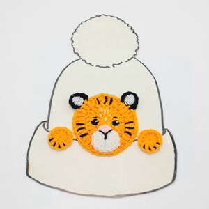 May include: A white crocheted hat with a pom-pom on top. A crocheted orange and black tiger face is attached to the front of the hat.