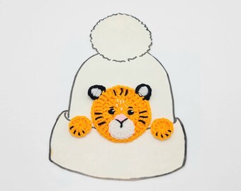 May include: A white crocheted hat with a pom-pom on top. A crocheted orange and black tiger face is attached to the front of the hat.