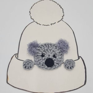 Puede incluir: Un gorro blanco de punto con un pomp&oacute;n esponjoso en la parte superior. Un koala de punto gris est&aacute; sujeto a la parte delantera del gorro.