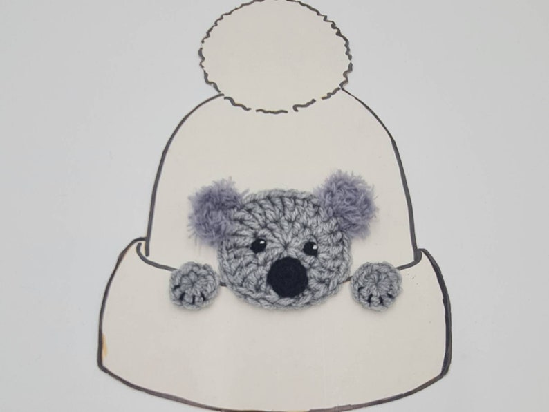 Puede incluir: Un gorro blanco de punto con un pomp&oacute;n esponjoso en la parte superior. Un koala de punto gris est&aacute; sujeto a la parte delantera del gorro.