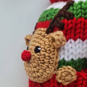 May include: A red, green, and white knitted hat with a brown reindeer face and antlers. The reindeer has a red nose and black eyes.
