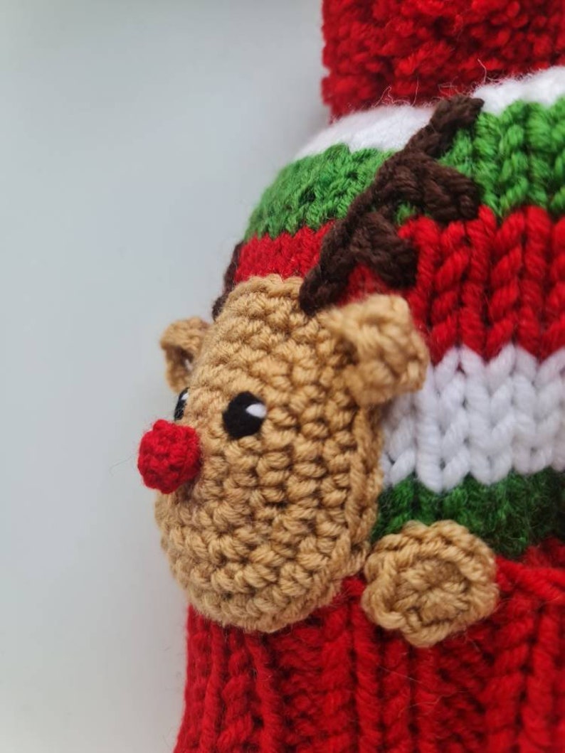 May include: A red, green, and white knitted hat with a brown reindeer face and antlers. The reindeer has a red nose and black eyes.