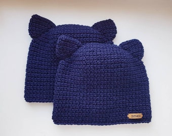May include: Two navy blue crocheted hats with ear flaps. The hats are made with a textured stitch and have a brown leather tag with the word "21nice" on it.