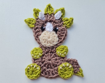 May include: A brown crocheted dinosaur with green leaves for a crest and green feet. The dinosaur has white eyes and a white nose.