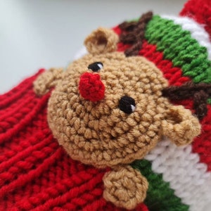 May include: A red, white, and green knitted hat with a brown crocheted reindeer face. The reindeer has a red nose and black eyes.