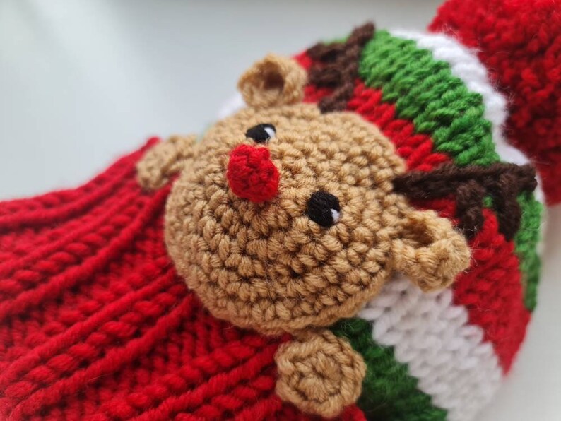 May include: A red, white, and green knitted hat with a brown crocheted reindeer face. The reindeer has a red nose and black eyes.
