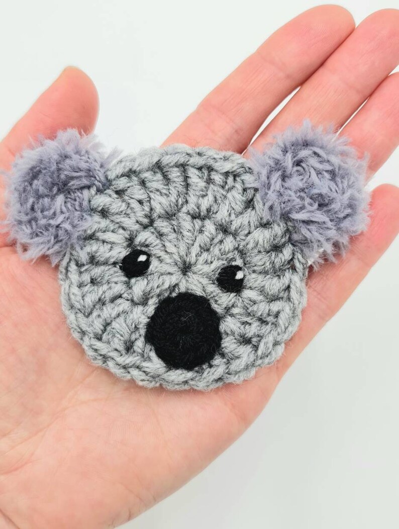 Puede incluir: Un koala de crochet gris con ojos y nariz negros. El koala tiene orejas grises y peludas.