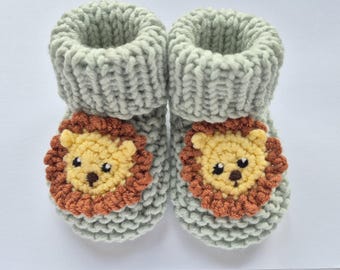 May include: A pair of light sage green knitted booties featuring a textured cuff and a crocheted lion face appliqué on the front of each shoe. The lion faces are crafted with yellow yarn, brown manes, and dark brown embroidered eyes and noses.