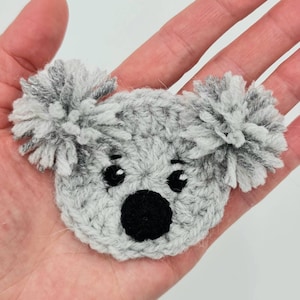 May include: A grey crocheted koala bear applique with black eyes and a black nose. The koala has fluffy grey ears.