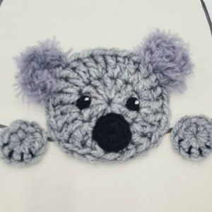 Puede incluir: Un oso koala de crochet gris con ojos y nariz negros, asom&aacute;ndose desde detr&aacute;s de una superficie blanca. El koala tiene orejas y patas grises y peludas.