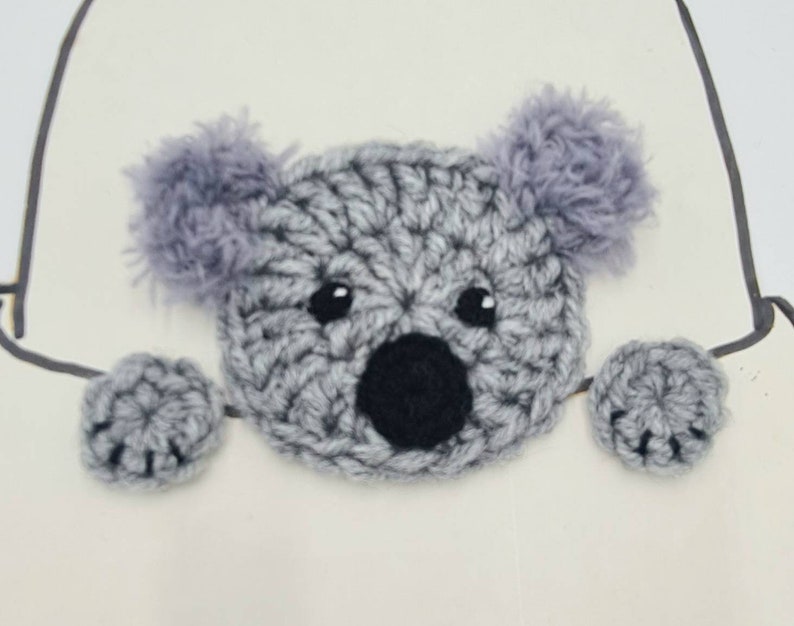 Puede incluir: Un oso koala de crochet gris con ojos y nariz negros, asom&aacute;ndose desde detr&aacute;s de una superficie blanca. El koala tiene orejas y patas grises y peludas.