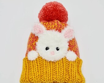 May include: A knitted beanie hat with a bunny face design. The hat features a yellow brim, a white bunny face with pink ears, and a red pom-pom on top. The body of the hat is orange and yellow.