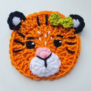 May include: A crocheted tiger face with orange fur, black stripes, white whiskers, and a pink nose. The tiger is wearing a green bow.