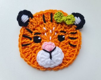 May include: A crocheted tiger face with orange fur, black stripes, white whiskers, and a pink nose. The tiger is wearing a green bow.