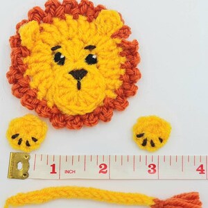 May include: A crocheted lion applique with a yellow body, orange mane, and black eyes. The lion is made up of three pieces: the head, a tail, and two paws. The pieces are laid out on a white surface with a measuring tape.