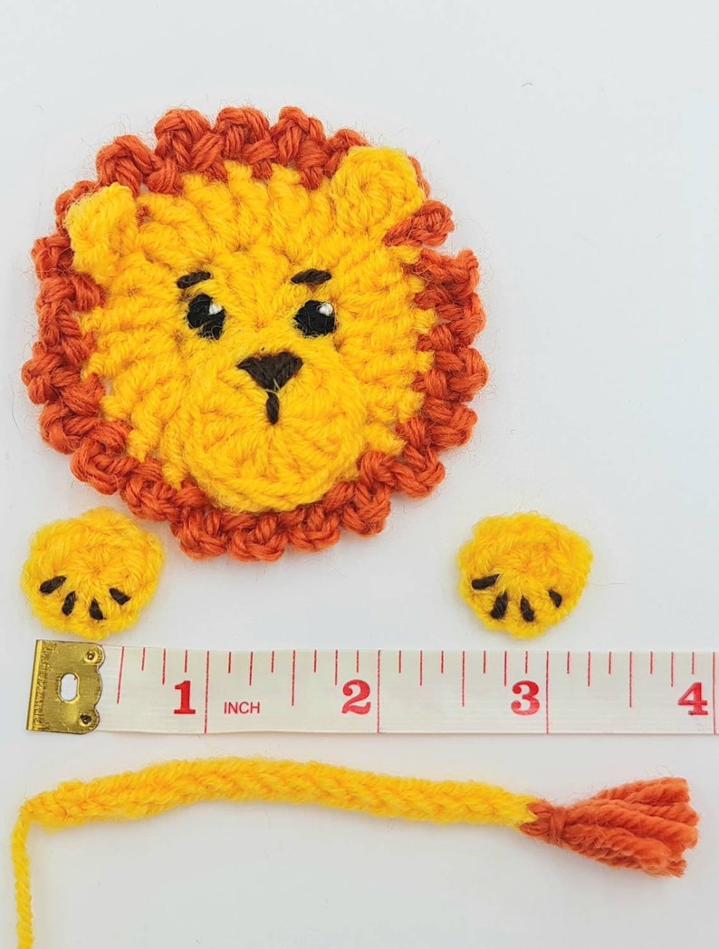 May include: A crocheted lion applique with a yellow body, orange mane, and black eyes. The lion is made up of three pieces: the head, a tail, and two paws. The pieces are laid out on a white surface with a measuring tape.