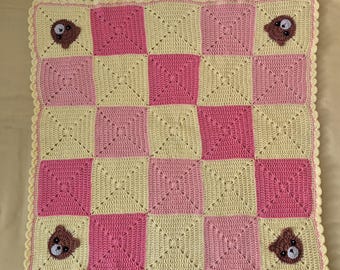 May include: A square crochet blanket featuring a checkerboard pattern of pale yellow and bright pink squares. The four corner squares each contain a small, brown, crocheted bear face with black eyes and a white muzzle. The edge has a scalloped border.