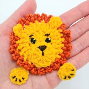 May include: A crocheted lion applique with a yellow face, orange mane, and black eyes. The lion has two yellow paws with black claws.
