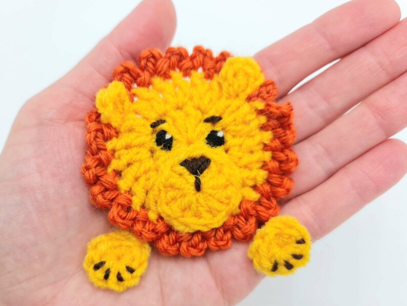 May include: A crocheted lion applique with a yellow face, orange mane, and black eyes. The lion has two yellow paws with black claws.