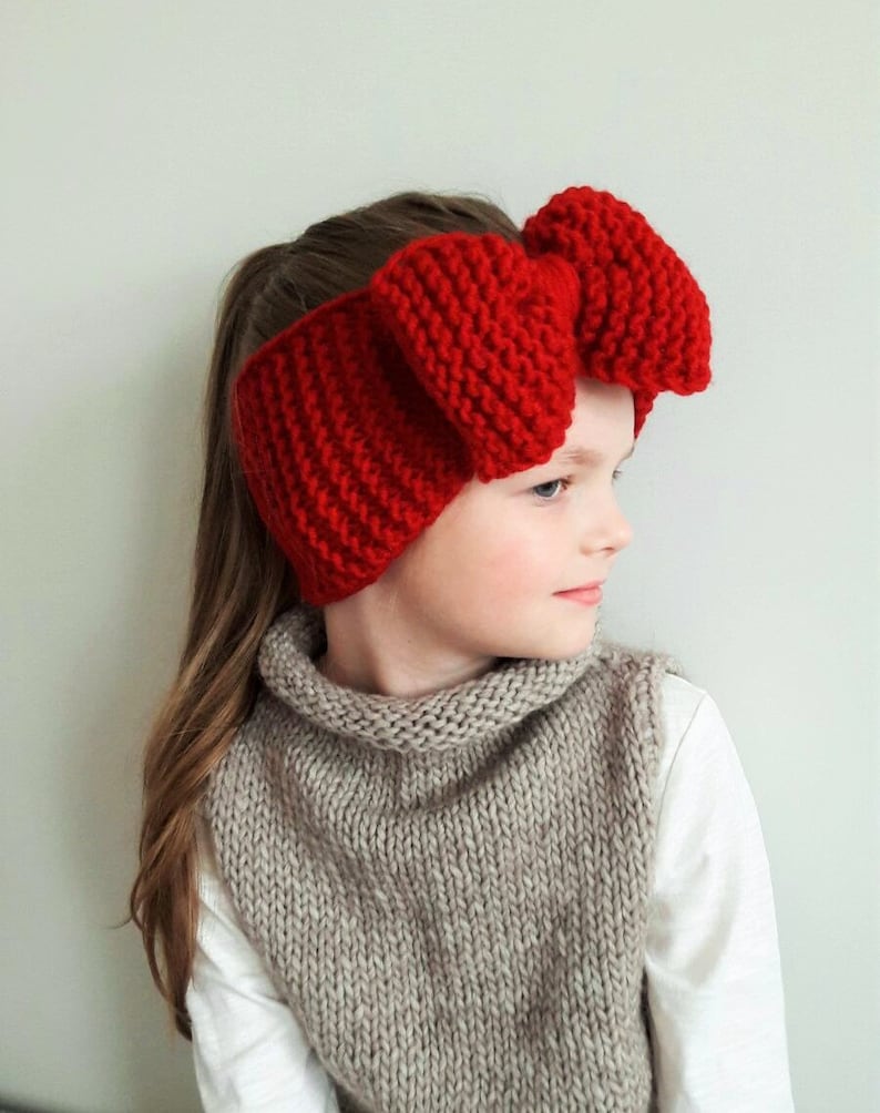 May include: A red knitted headband with a large bow. The headband is worn by a young girl with long brown hair.