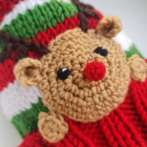 May include: A red, green, and white striped knitted hat with a brown reindeer face attached. The reindeer has a red nose and black eyes.