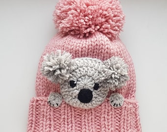 May include: Pink knitted hat with a fluffy pom pom on top and a gray knitted koala face on the front.
