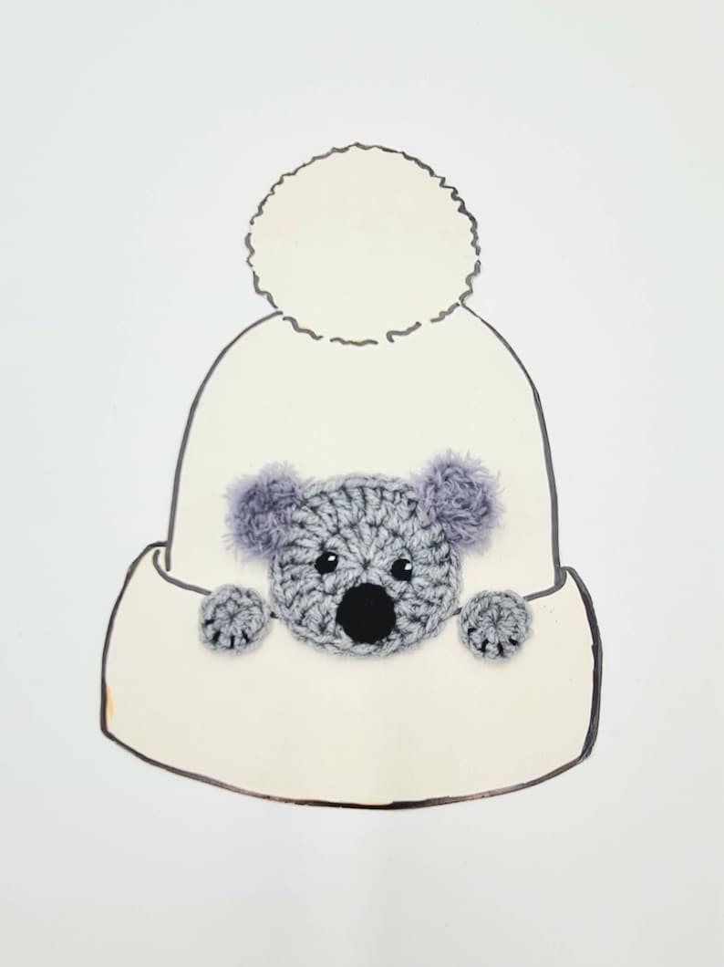 Puede incluir: Un gorro blanco de punto con un pomp&oacute;n esponjoso en la parte superior. Un koala de punto gris asoma desde el gorro.