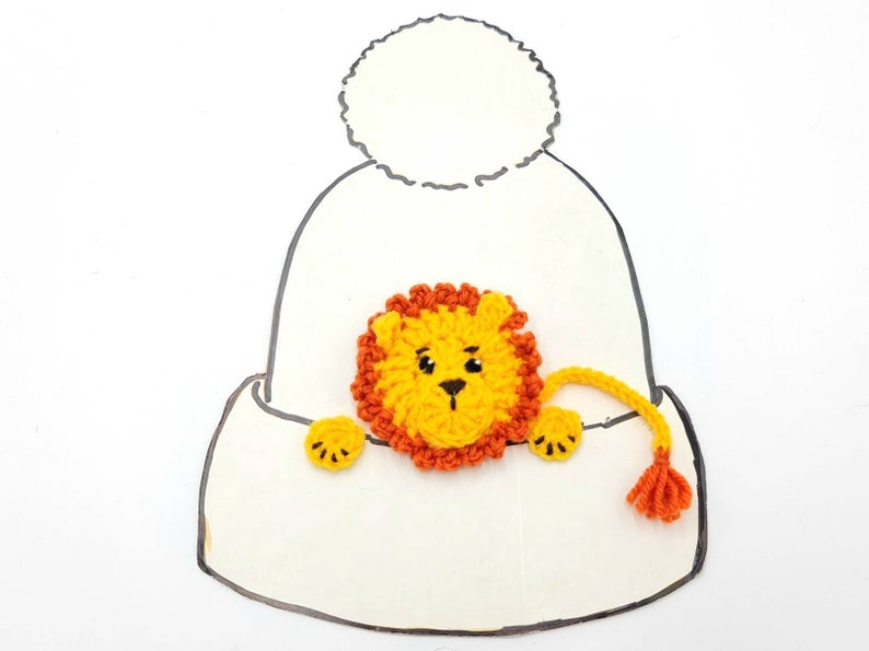May include: A white crocheted hat with a pom-pom on top. A yellow crocheted lion with an orange mane is peeking out of the hat.