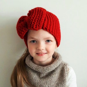 May include: A young girl wearing a red knitted headband with a large bow on top. The headband is made with a chunky yarn and has a simple, classic design.