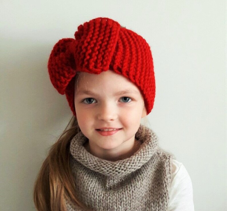May include: A young girl wearing a red knitted headband with a large bow on top. The headband is made with a chunky yarn and has a simple, classic design.