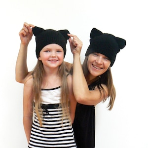 May include: Two people wearing black knit hats with cat ears. The woman is wearing a black dress and the girl is wearing a black and white striped dress.