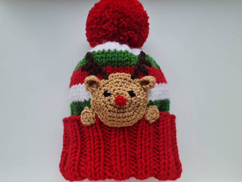 May include: Red and green striped knit hat with a white stripe and a brown reindeer face with a red nose. The hat has a red pom-pom on top.