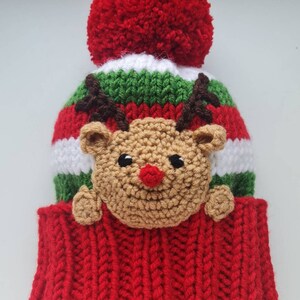 May include: Red and green knitted hat with a white stripe and a brown reindeer applique. The hat has a red pom pom on top.