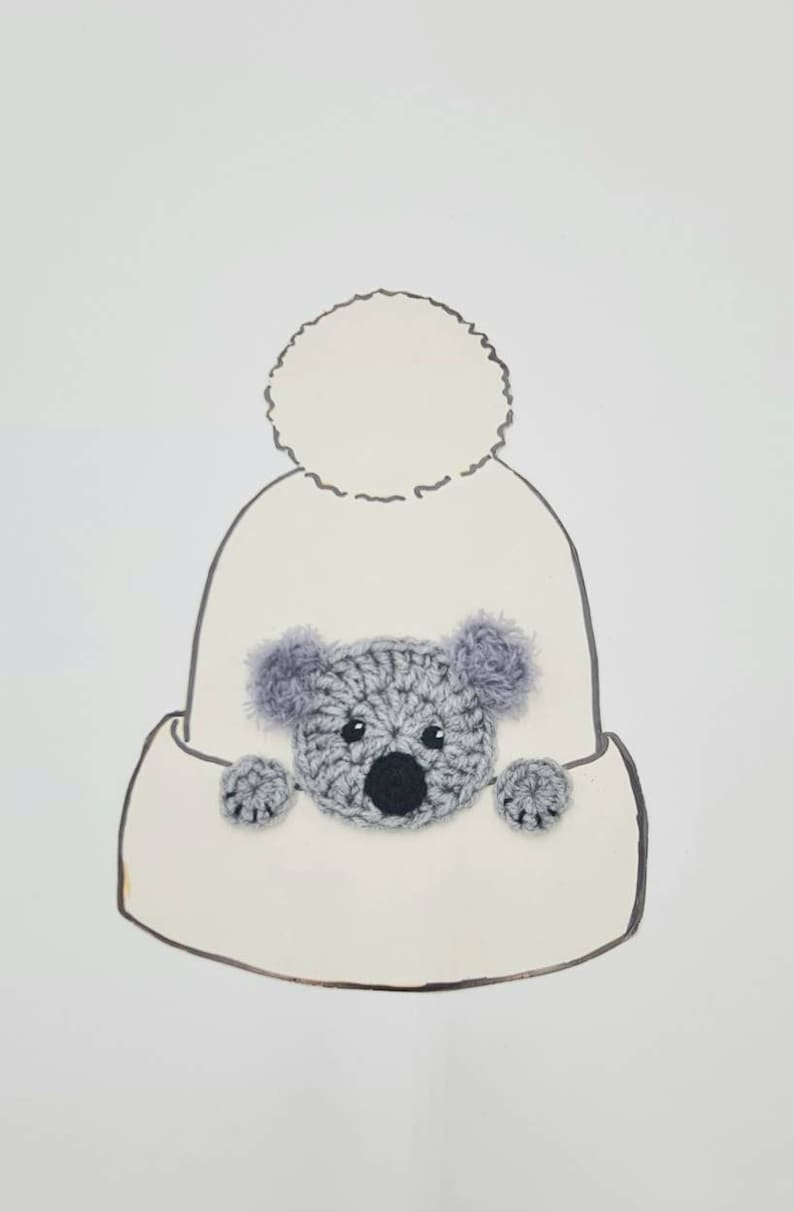 Puede incluir: Gorro blanco de punto con un pomp&oacute;n esponjoso en la parte superior y una aplicaci&oacute;n de koala de punto gris en la parte delantera.
