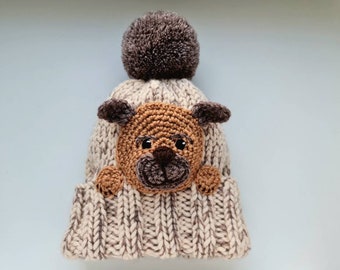 May include: A beige knitted hat with a brown pom pom on top and a brown crocheted dog face appliqué.