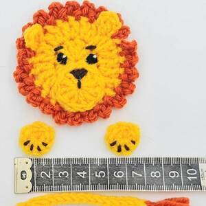 May include: Crocheted lion applique with a yellow body, orange mane, and black eyes. The lion has two small yellow paws with black claws and a long yellow tail with an orange tip. The lion is measured against a ruler.
