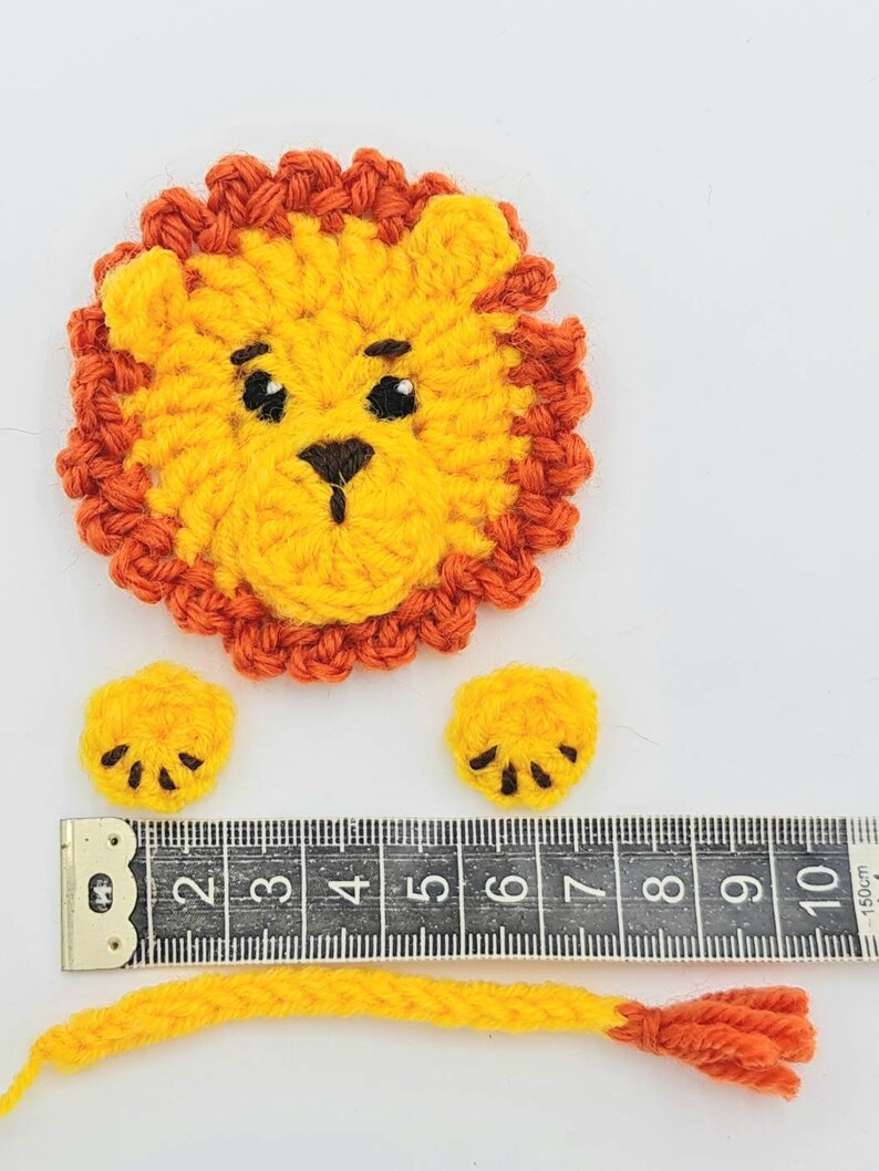 May include: Crocheted lion applique with a yellow body, orange mane, and black eyes. The lion has two small yellow paws with black claws and a long yellow tail with an orange tip. The lion is measured against a ruler.