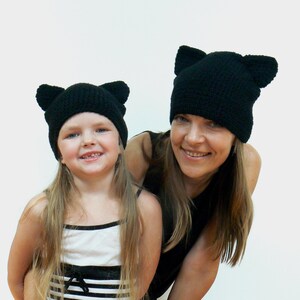 May include: Two people wearing black knit hats with cat ears. The hats are crocheted and have a simple design. The woman is wearing a black top and the girl is wearing a striped tank top.