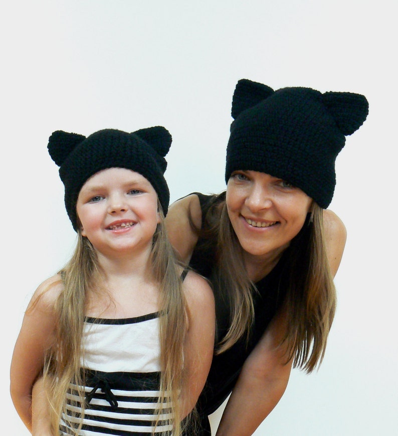May include: Two people wearing black knit hats with cat ears. The hats are crocheted and have a simple design. The woman is wearing a black top and the girl is wearing a striped tank top.
