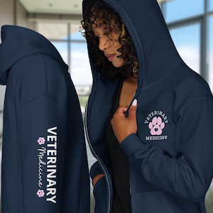 May include: A dark navy blue zip-up hoodie featuring a small pink paw print logo with the text Veterinary Medicine on the chest. The sleeve displays the text Veterinary Medicine vertically in white, accented by two small pink paw prints.