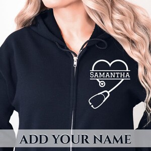 May include: A black zip-up hoodie featuring a white graphic on the left chest. The design consists of a heart shape integrated with a stethoscope and the name SAMANTHA printed in the centre. The hoodie includes a hood with black drawstrings.