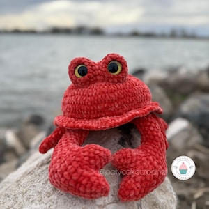 Pode incluir: Um caranguejo de crochê vermelho com olhos amarelos sentado em uma rocha. O caranguejo é feito com um fio macio e felpudo e tem uma expressão fofa e amigável. O caranguejo está sentado em uma rocha perto da água.