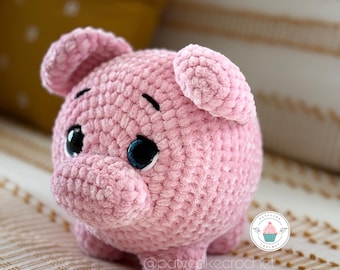May include: A pink crocheted pig stuffed animal with black eyes and a small, pink snout. The pig is sitting on a white and gold striped surface.